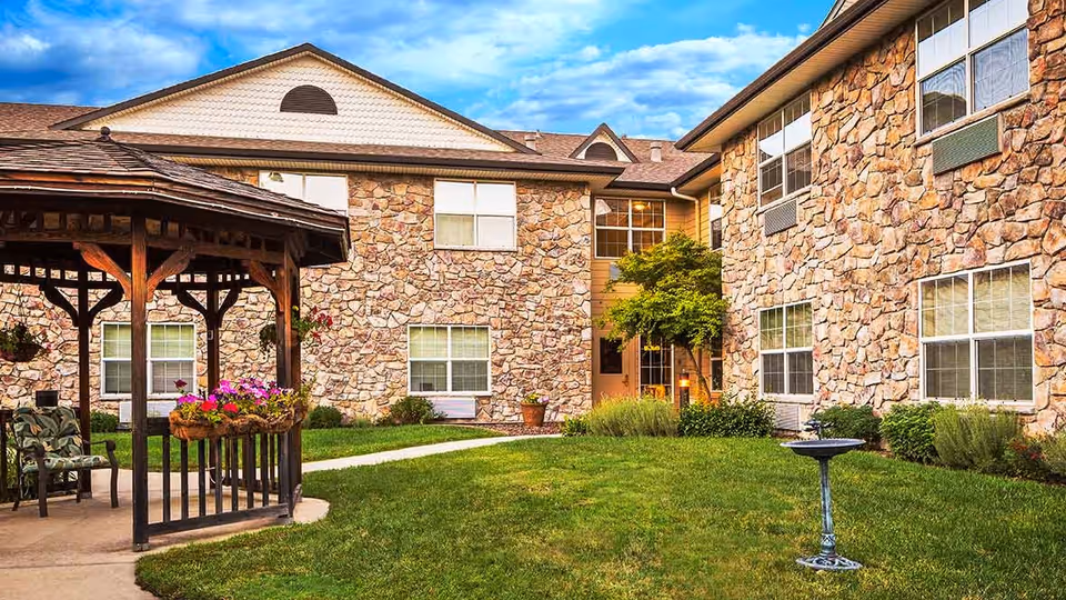 Outdoor courtyard area of a senior living facility with stone exterior walls, a wooden gazebo with hanging flower baskets, green grass, a birdbath, and a small tree near the building entrance.