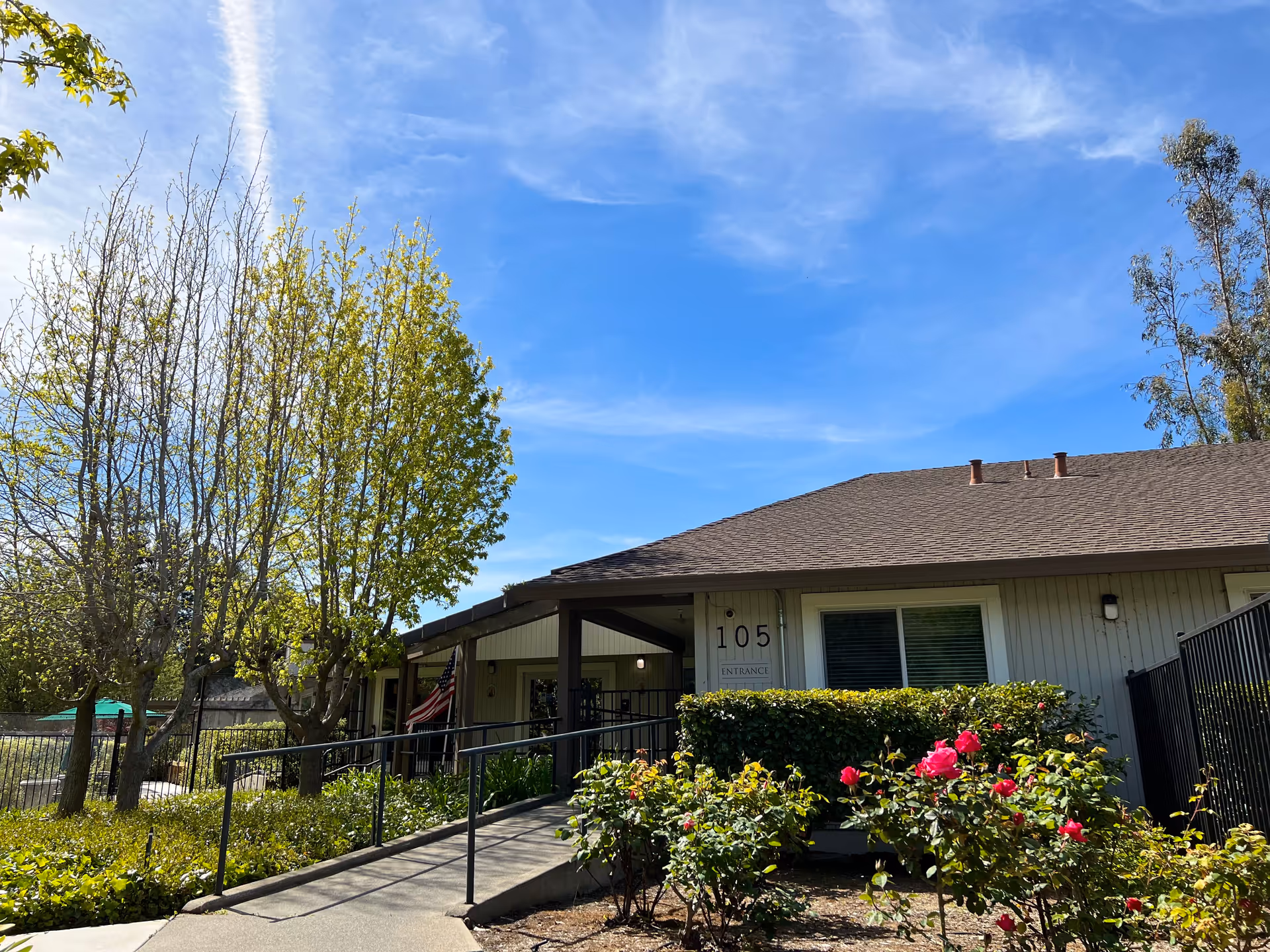 Exterior front of a single-story Vista Prado building marked '105' with a ramp, shrubs and blooming roses under a blue sky.