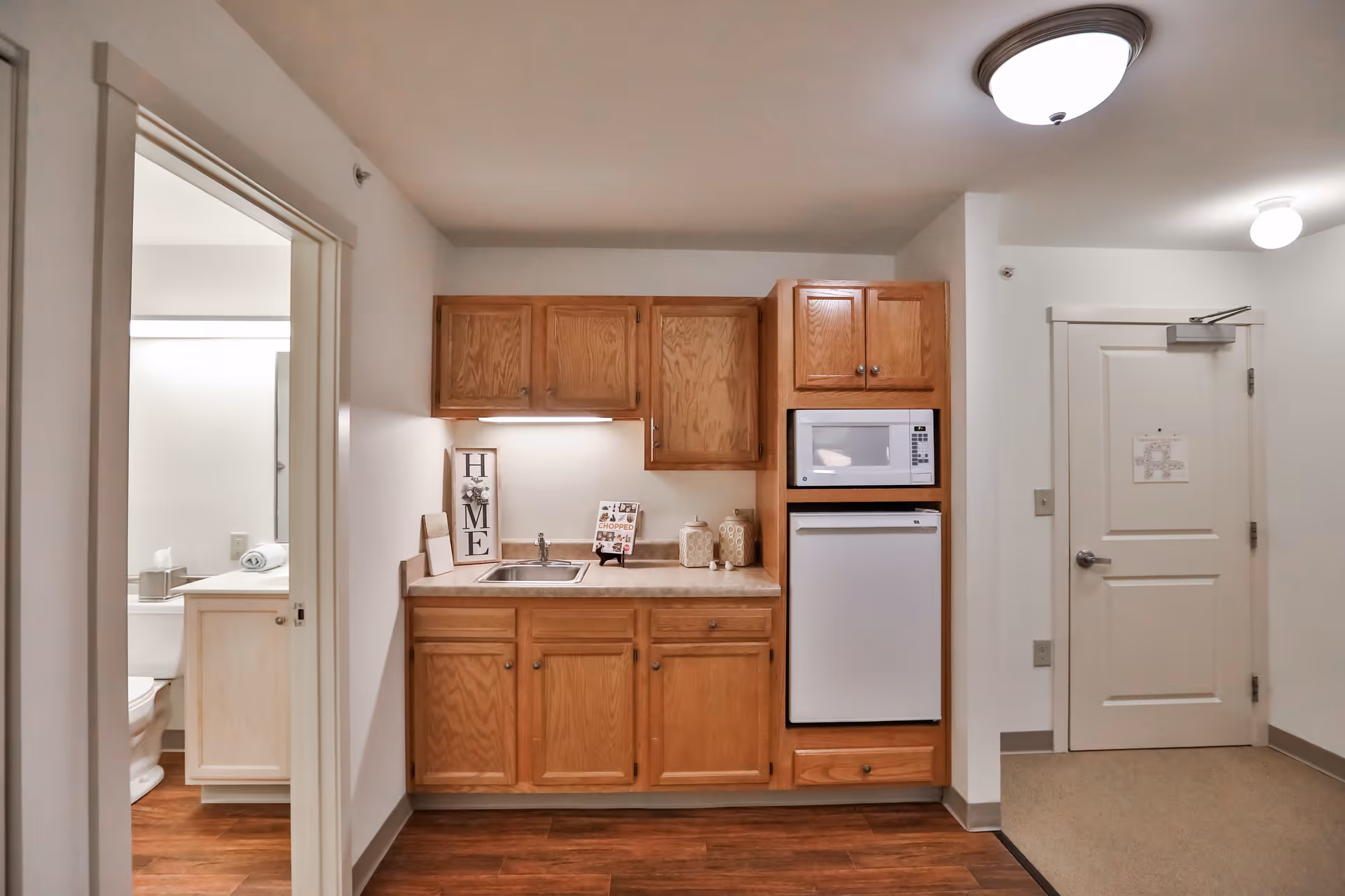 Interior view of a small kitchen area with wooden cabinets, a sink, a microwave, and a mini refrigerator. To the left, there is an open doorway leading to a bathroom with a toilet and a sink visible. The floor is wood laminate, and the walls are painted white. A closed white door is on the right side of the kitchen area.