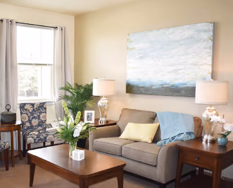 A cozy living room with a beige sofa adorned with a yellow pillow and a blue throw blanket. Two wooden side tables with glass lamps flank the sofa. A wooden coffee table with a vase of white flowers sits in front of the sofa. A patterned armchair is positioned near a window with light curtains. A large abstract painting hangs on the wall above the sofa.