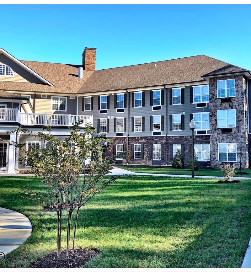 Exterior view of a multi-story senior living facility building with beige and stone facade, multiple windows, a chimney, and a balcony. The foreground features a well-maintained green lawn with small trees and a paved walkway under a clear blue sky.