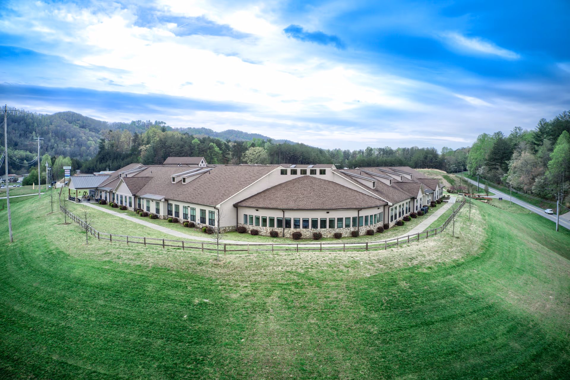 Wide exterior view of a single-story assisted living facility with a brown roof and beige walls, surrounded by green grass and trees, under a partly cloudy sky.