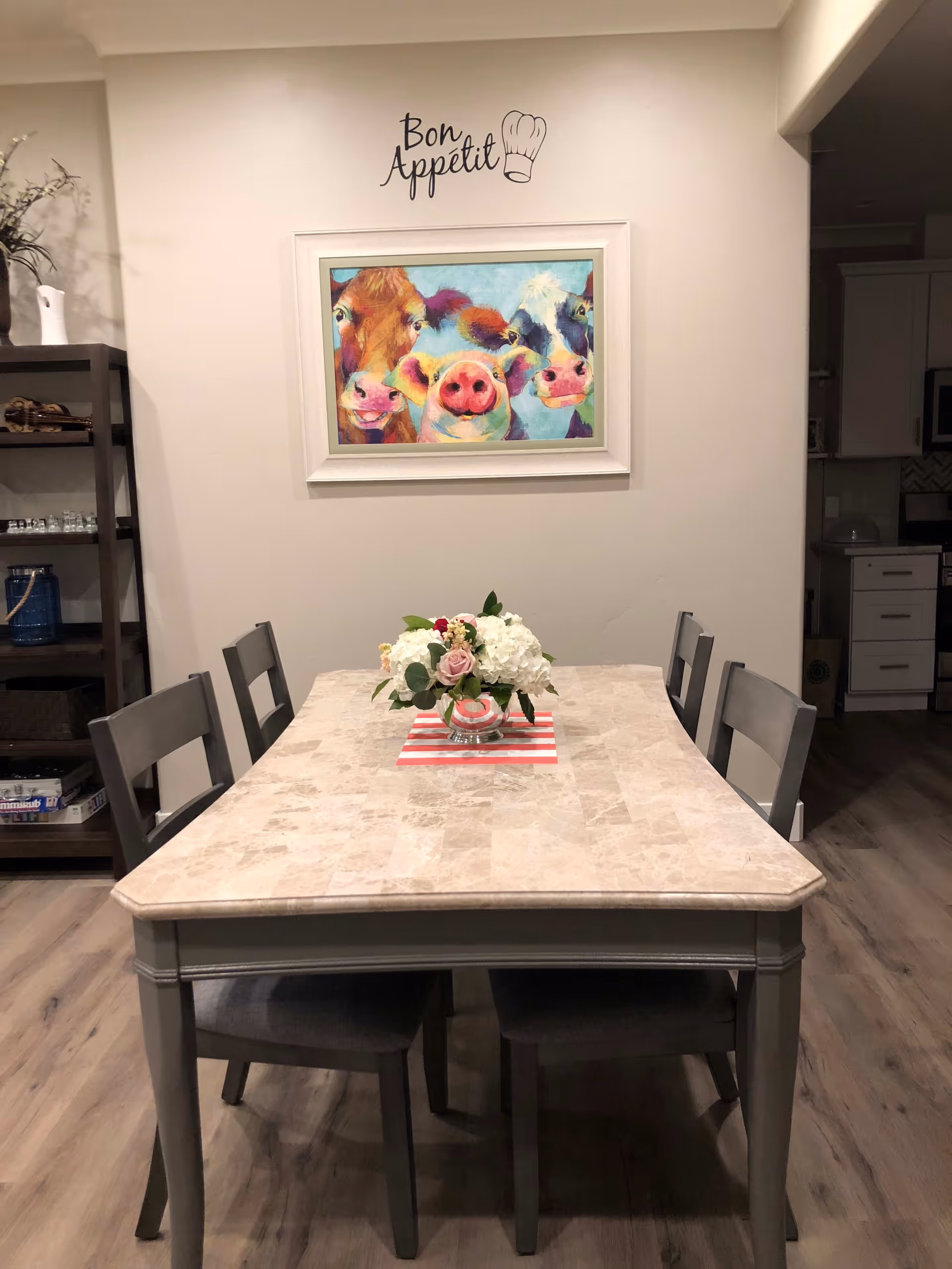 A dining area with a rectangular table and four chairs. The table has a light-colored stone top and a floral centerpiece with white and pink flowers on a red and white striped placemat. On the wall above the table is a colorful painting of three farm animals: a cow, a pig, and another cow. Above the painting, the words 'Bon Appétit' and a chef's hat illustration are displayed. To the left, there is a dark wooden shelving unit with various items, and to the right, part of a kitchen area is visible.