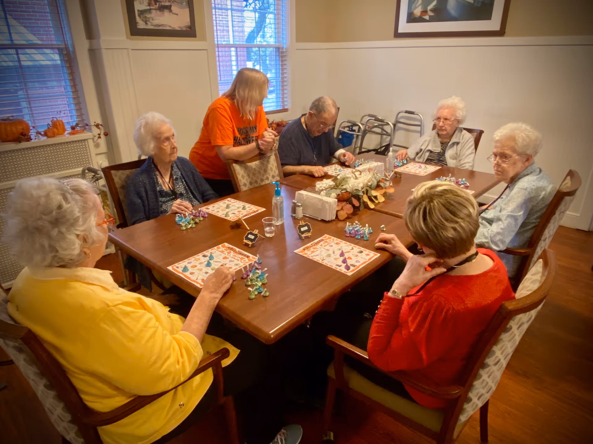 A group of elderly people sitting around a wooden table playing a board game in a well-lit room with windows and framed pictures on the walls. A caregiver in an orange shirt is standing and assisting one of the players.