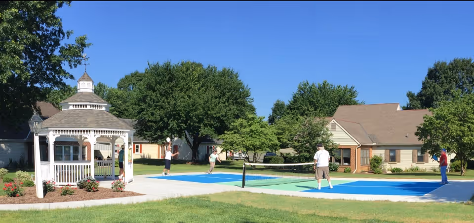 Outdoor scene at Homestead Village showing a group of elderly people playing pickleball on a blue and green court. There is a white gazebo on the left side surrounded by flower beds, with trees and residential buildings in the background under a clear blue sky.