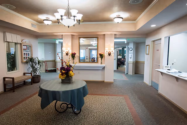 A senior living facility lobby with a round table topped with a floral arrangement, chandelier lighting, seating, and a reception window.