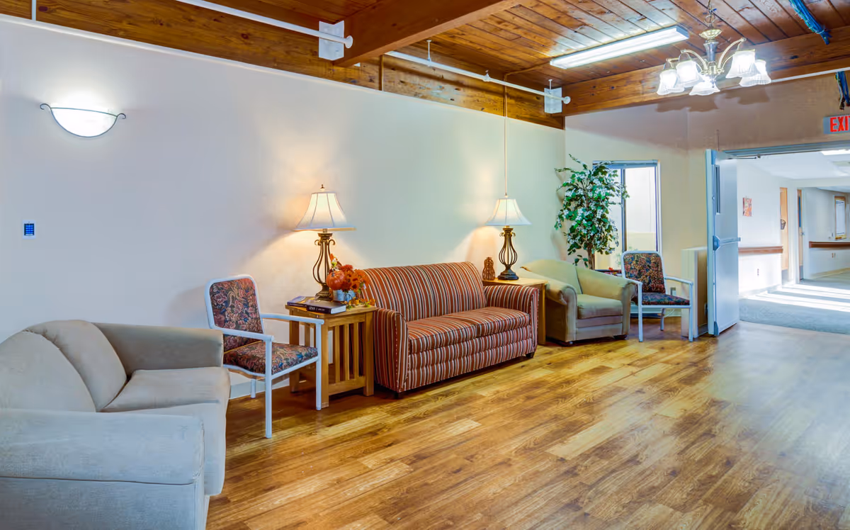 A cozy seating area in a senior living facility with a beige couch, a striped sofa, two floral-patterned chairs, two wooden side tables with lamps, a potted plant, and wooden flooring under a wooden beam ceiling with overhead lighting.