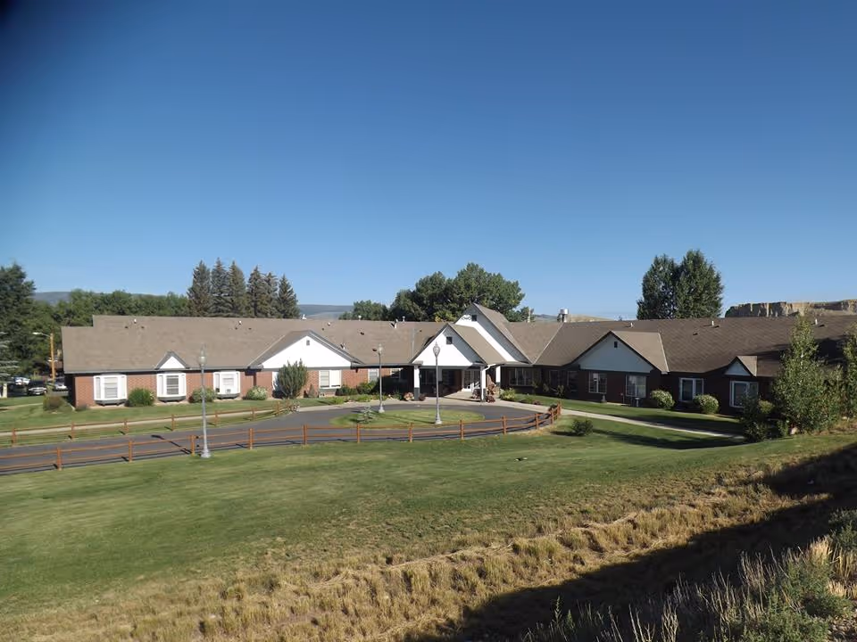 Front exterior of a single-story brick assisted living building with a circular driveway and well-kept lawn under a clear blue sky.