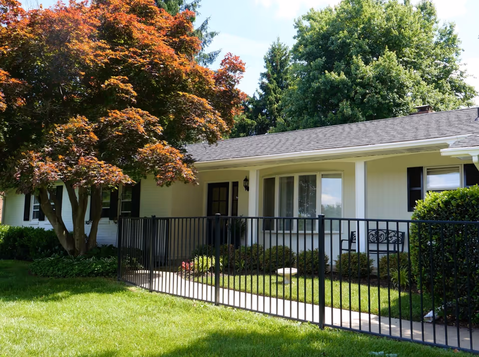 Exterior view of a single-story building with a black metal fence in front, a concrete walkway, green grass, and a large tree with reddish leaves on the left side. The building has light-colored walls, black shutters, and a small porch area with a bench.