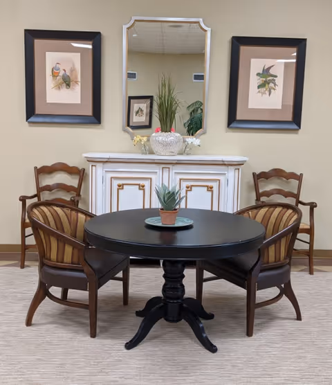 A small seating area with a round black table surrounded by four wooden chairs with striped cushions. Behind the table is a white cabinet with gold accents, topped with a decorative vase and flowers. Above the cabinet is a rectangular mirror flanked by two framed bird illustrations on a beige wall.