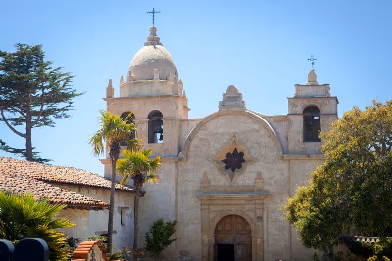 Exterior view of a historic mission-style building with a dome, bell towers, and a cross on top, surrounded by palm trees and other greenery under a clear blue sky.