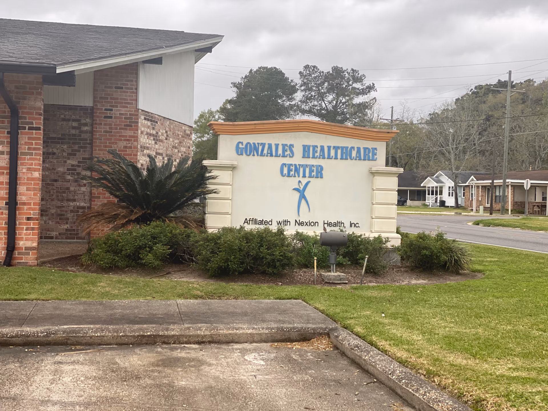 Outdoor view of the Gonzales Healthcare Center sign next to a brick building with greenery and a street in the background under a cloudy sky.