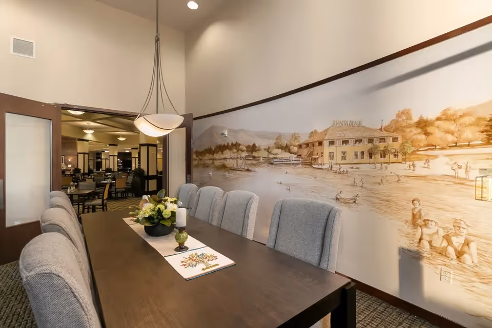 A dining room with a long dark wooden table surrounded by gray upholstered chairs. A decorative centerpiece with flowers and candles is on the table. The wall features a large mural depicting a vintage beach scene with people swimming and a building labeled 'Juanita Beach'. Double doors open to another room with more tables and chairs.