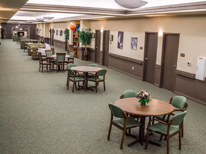 A spacious interior hallway in a senior living facility with round tables and green cushioned chairs arranged along the carpeted corridor. There are doors along the right wall, framed pictures, potted plants, and comfortable seating areas further down the hallway. The ceiling has recessed lighting and a large circular light fixture.