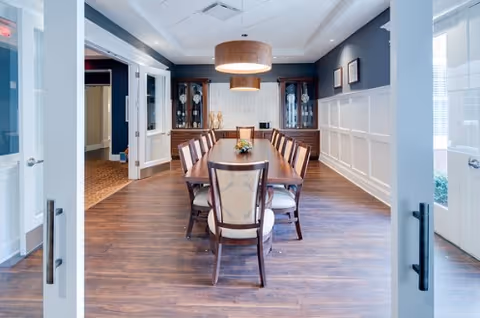 View through open double doors into a dining room with a long wooden table surrounded by chairs. The room features dark wood flooring, white wainscoting on the walls, two glass-front cabinets at the far end, and a large round pendant light hanging above the table.