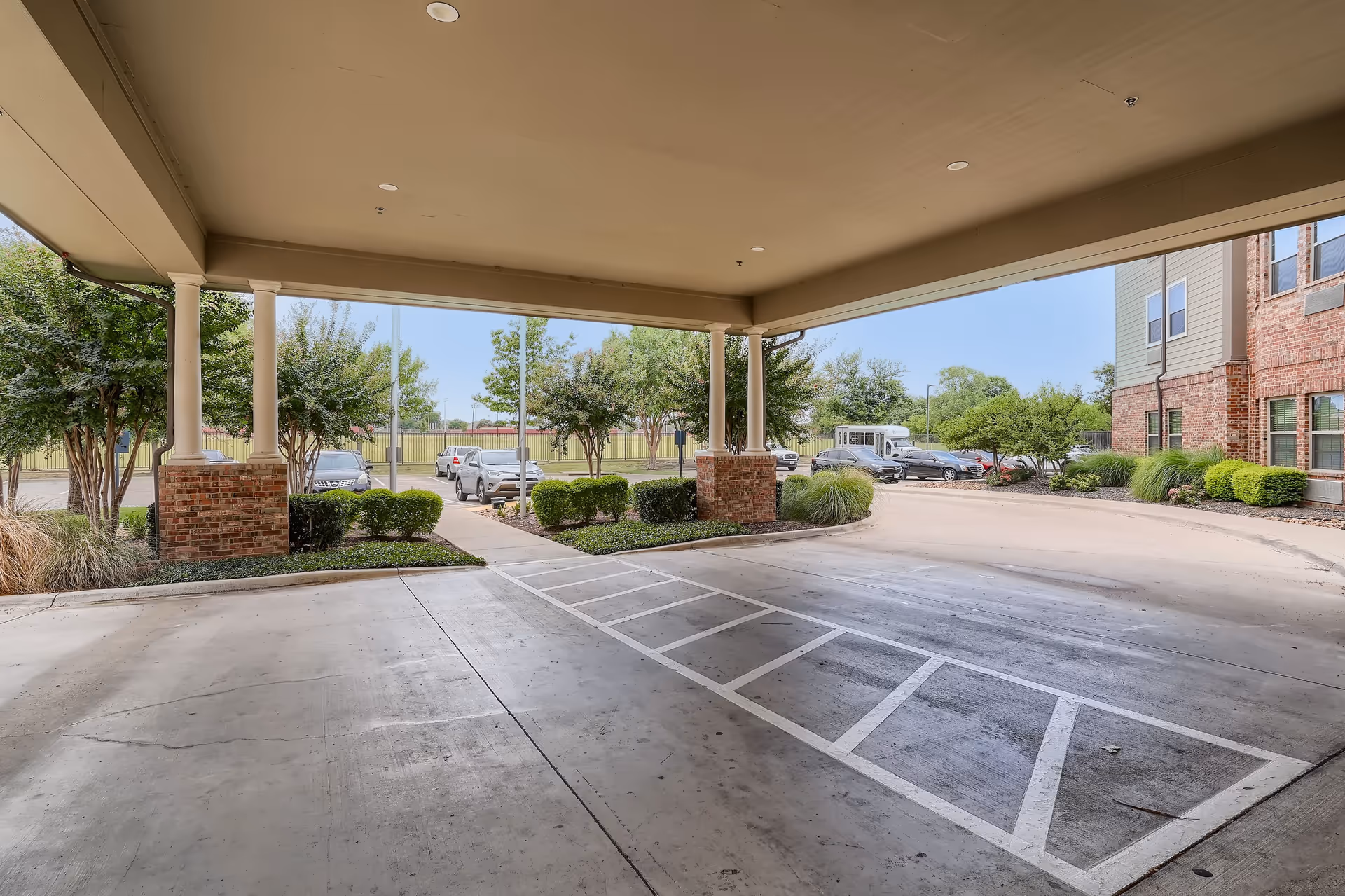Covered driveway entrance of a senior living facility with brick pillars and landscaped bushes, trees, and parked cars visible in the background under a clear blue sky.