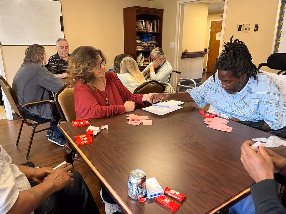 A group of elderly people sitting around a large wooden table in a common room, engaging in a card game. One woman in a red top is holding hands with a man in a hospital gown. There are several Kit Kat candy bars and a can of soda on the table. In the background, other residents are seated and interacting, with a bookshelf and a whiteboard visible.
