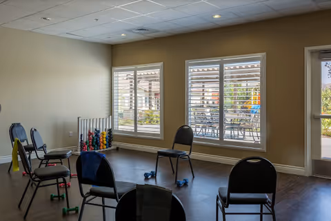 Bright interior activity room with chairs arranged in a circle, small dumbbells and exercise equipment by windows overlooking a patio.