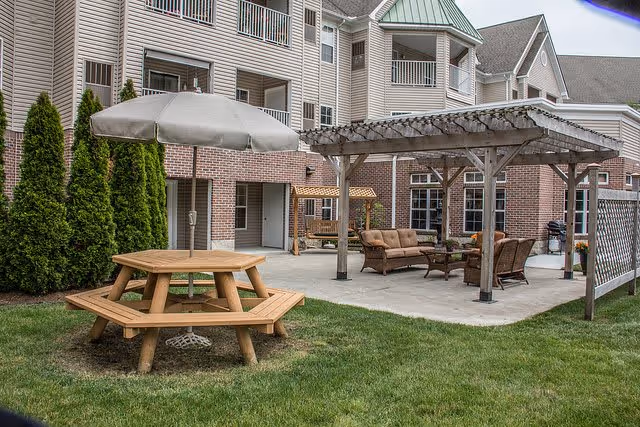 Outdoor patio area at The Village of Lake Huron Woods featuring a wooden picnic table with an umbrella, a pergola with cushioned seating including a sofa and chairs, and a brick building with balconies in the background.