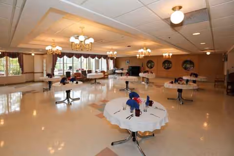 Bright spacious dining room with round tables, white tablecloths, centerpieces, and chandeliers.