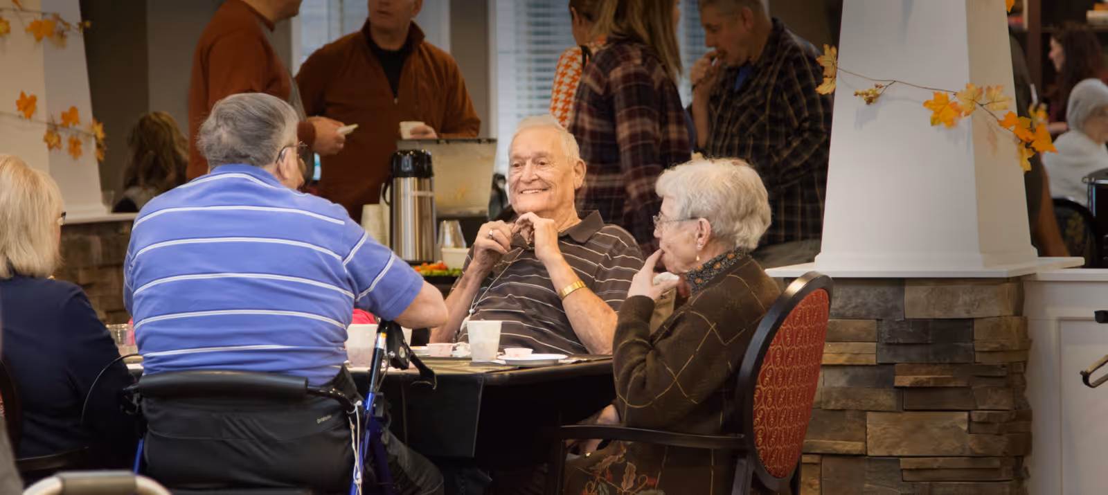 A group of elderly people sitting around a table in a communal dining or social area, engaging in conversation. The setting includes autumn-themed decorations with orange leaves on the walls and a stone pillar. Other people are visible in the background, standing and talking near a beverage station.
