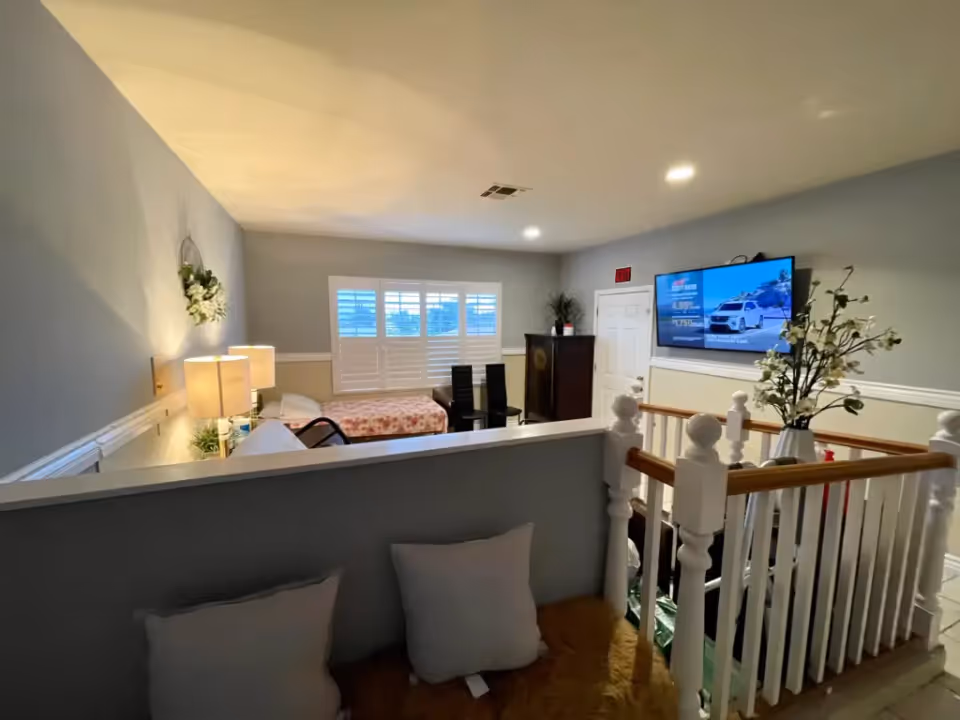 Open loft-style interior with a bed, seating, lamps, a wall-mounted TV, and a white staircase railing.
