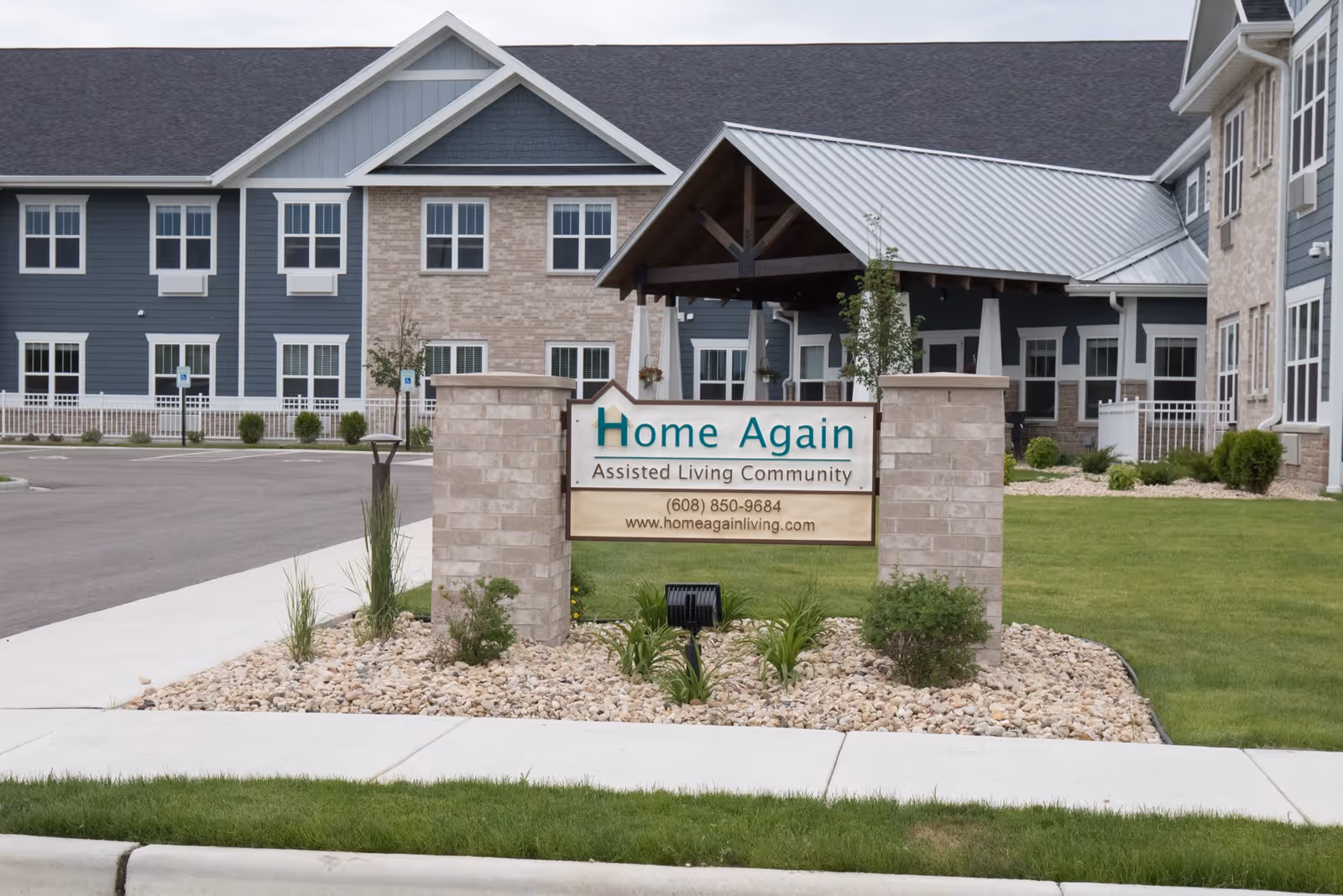 Exterior view of Home Again Assisted Living Community building with a sign in front displaying the facility name, phone number, and website. The building has blue and beige siding with multiple windows and a covered entrance. The surrounding area includes a paved driveway, green lawn, and landscaped rocks with plants.