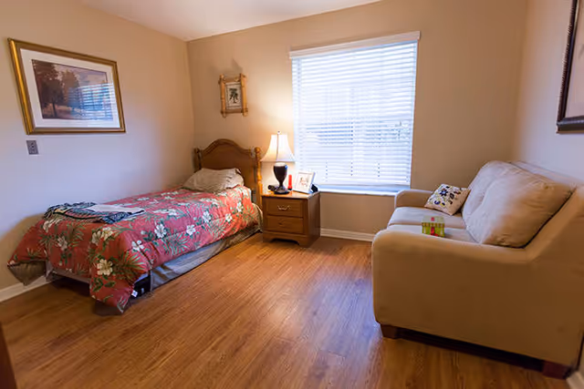 A cozy bedroom with a single bed covered in a red floral bedspread, a wooden nightstand with a lamp and framed photo, a window with white blinds, a beige couch with a decorative pillow, and framed artwork on the walls. The floor is wooden.