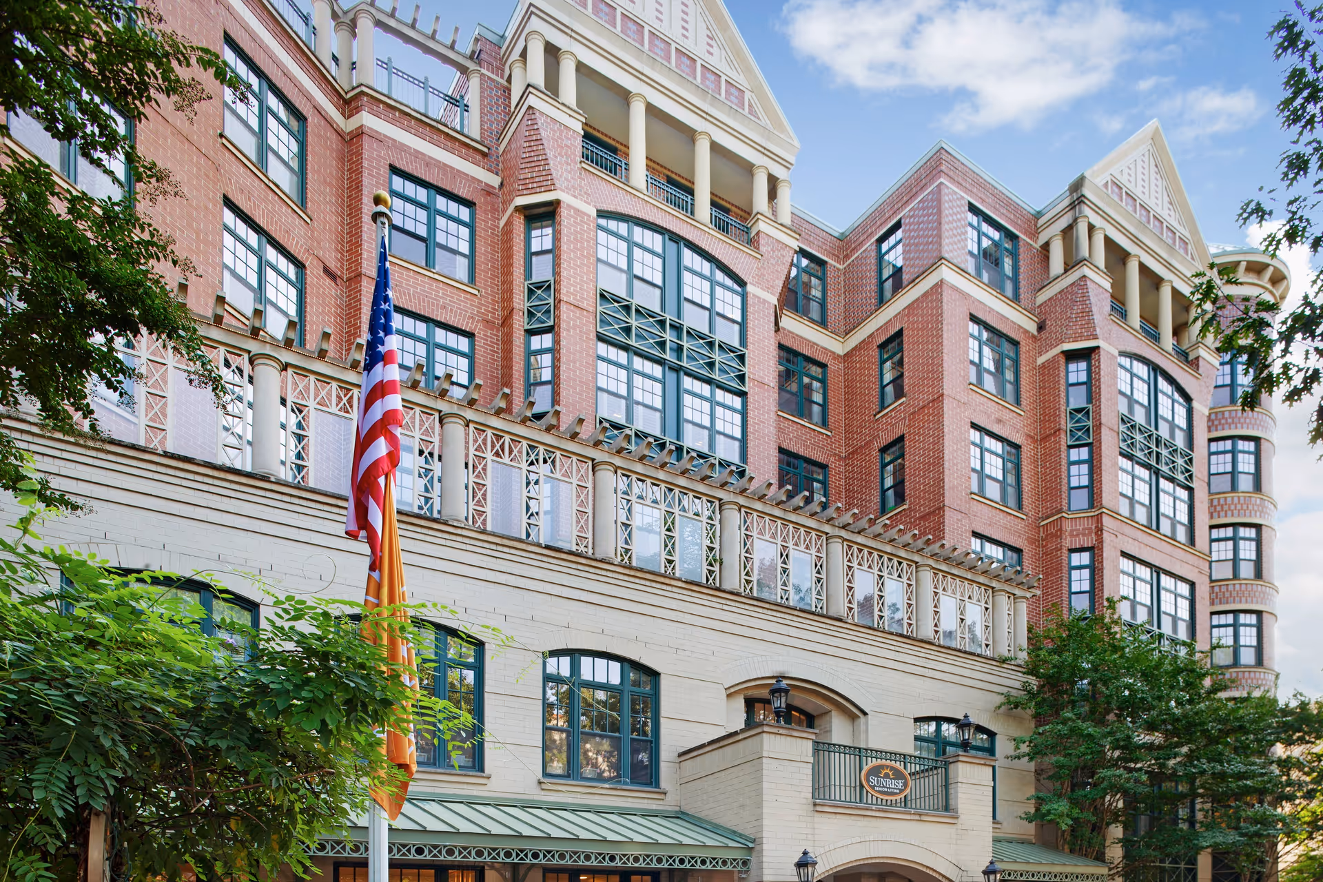 Front exterior of a multi-story red-brick senior living building with many windows, balconies, and an American flag in front.