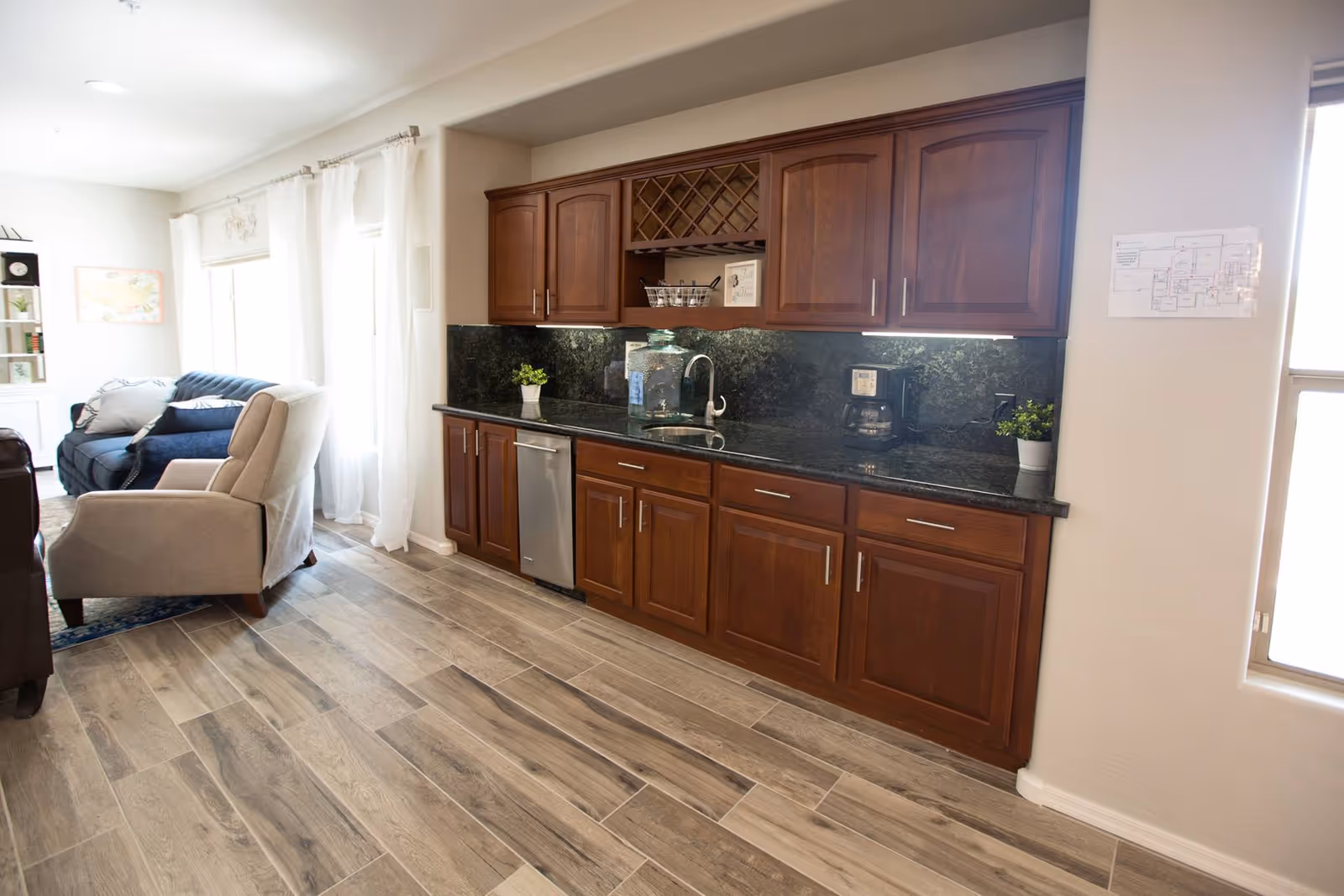 Interior view of a senior living facility showing a kitchenette area with dark wood cabinets, a granite countertop, a sink, a coffee maker, and small potted plants. Adjacent to the kitchenette is a living area with a beige armchair and a blue sofa near large windows with sheer white curtains allowing natural light to fill the room.