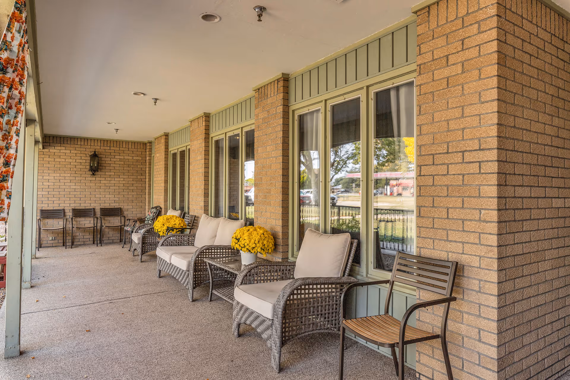 Covered outdoor patio area with wicker chairs and cushioned seats, small tables with yellow potted flowers, and additional chairs lined up against a brick wall under a ceiling with recessed lighting.