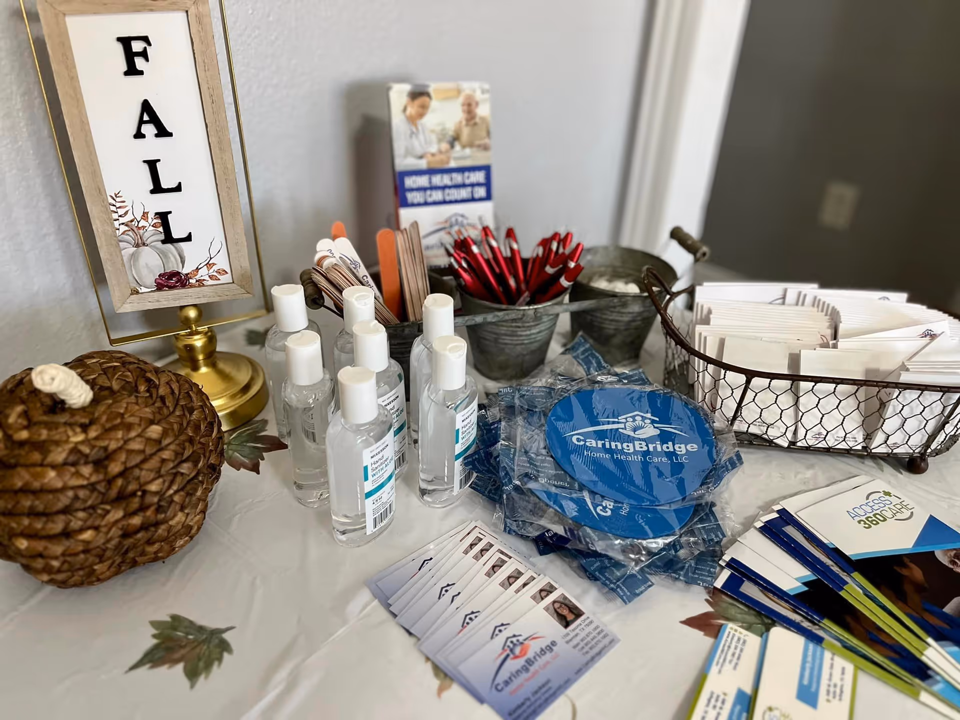 A table with various informational materials and promotional items including hand sanitizer bottles, pens, pamphlets, business cards, and a decorative sign that reads 'FALL'. There is also a woven basket shaped like a pumpkin and a metal basket holding folded papers.