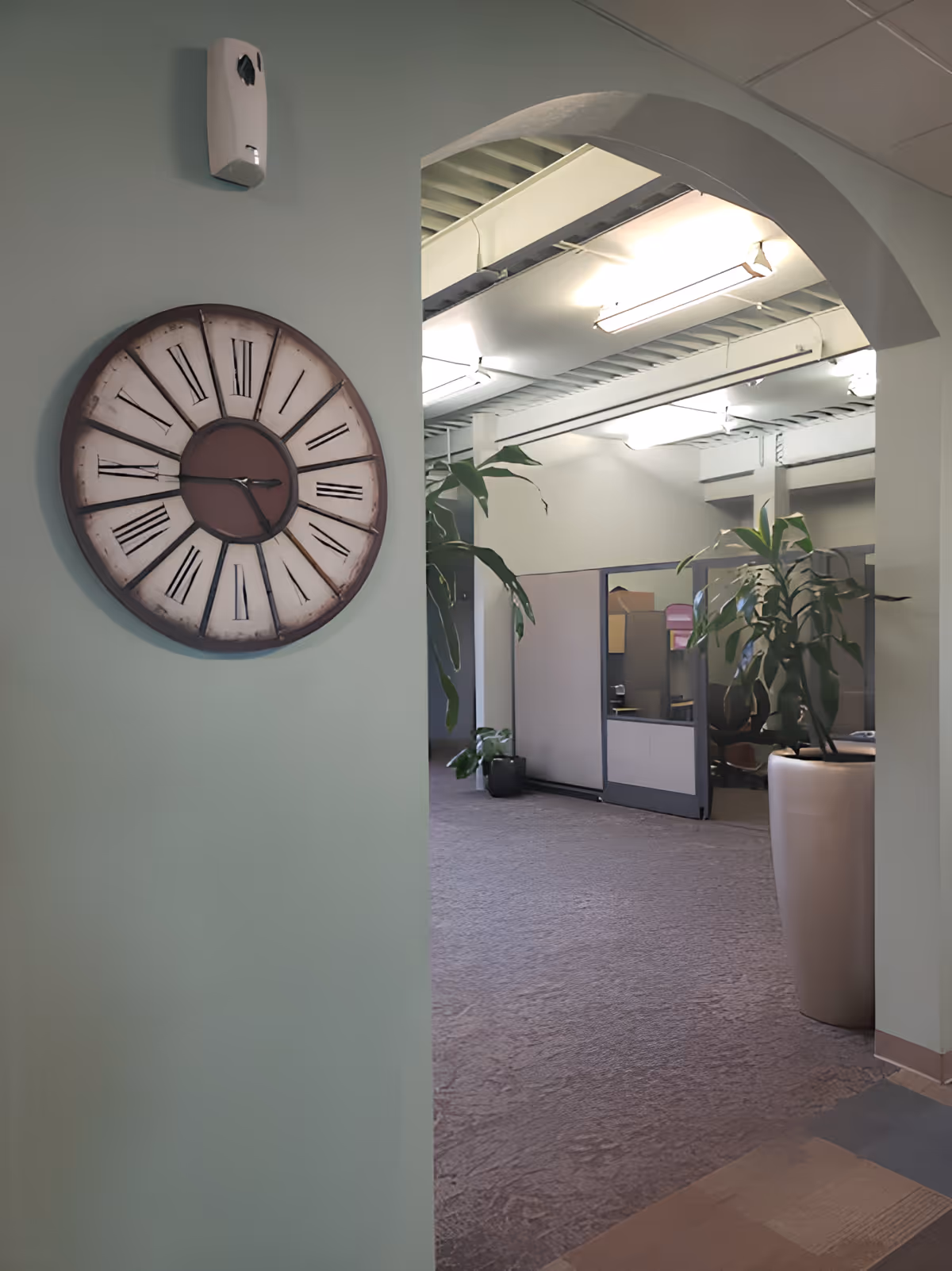 Interior view of a hallway in a facility with a large round wall clock showing the time as 3:45, a hand sanitizer dispenser mounted above it, and an arched doorway leading to an office area with cubicles and potted plants.