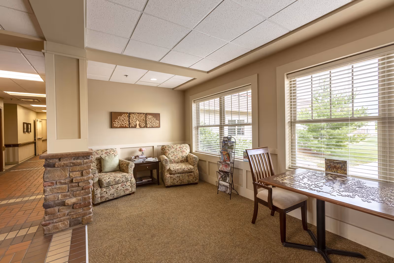 A cozy sitting area in a senior living facility with two floral armchairs and a small wooden side table between them. A wooden chair is placed next to a table with a partially completed jigsaw puzzle near large windows with blinds. There is a decorative tree artwork on the wall and a rack holding brochures or magazines near the window. The floor is carpeted and the ceiling has white tiles.