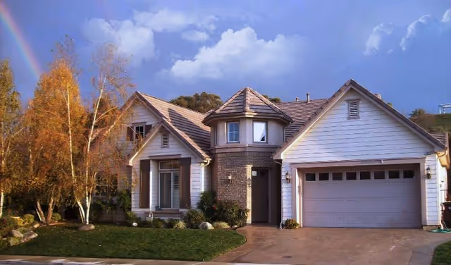 A single-story house with a two-car garage, a turret-like architectural feature, and a well-maintained front yard with green grass and trees. The sky is partly cloudy with a visible rainbow on the left side.
