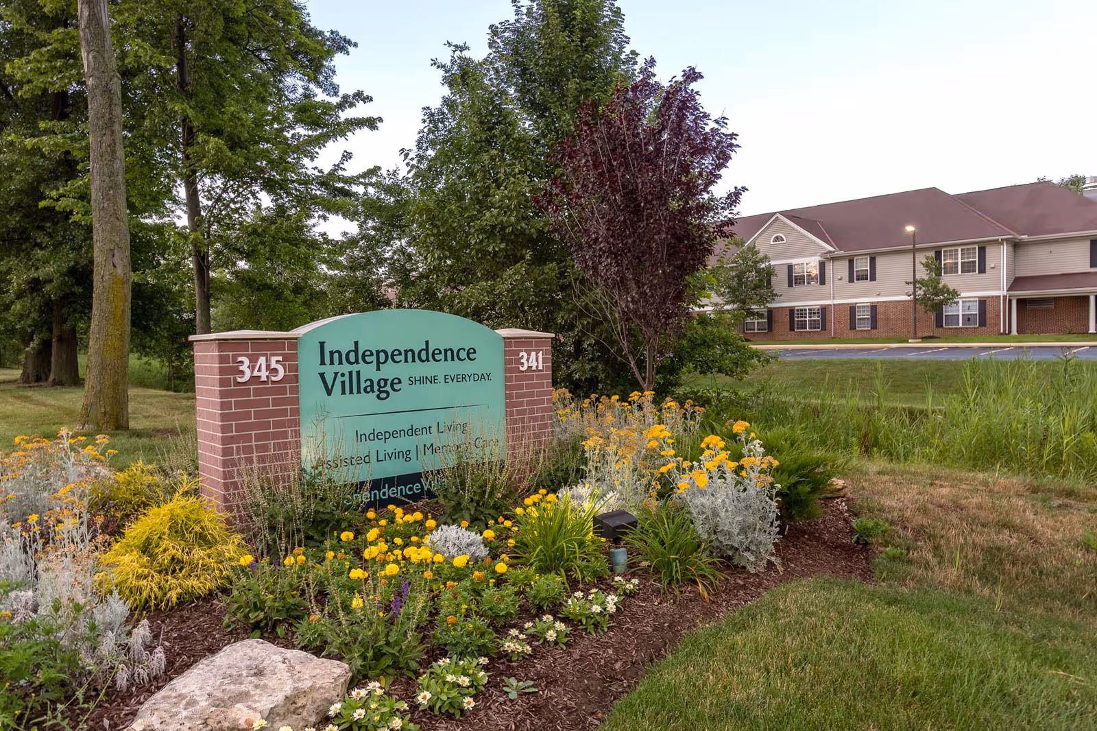 Landscaped entrance sign reading "Independence Village" with flowers in the foreground and the facility building behind it.