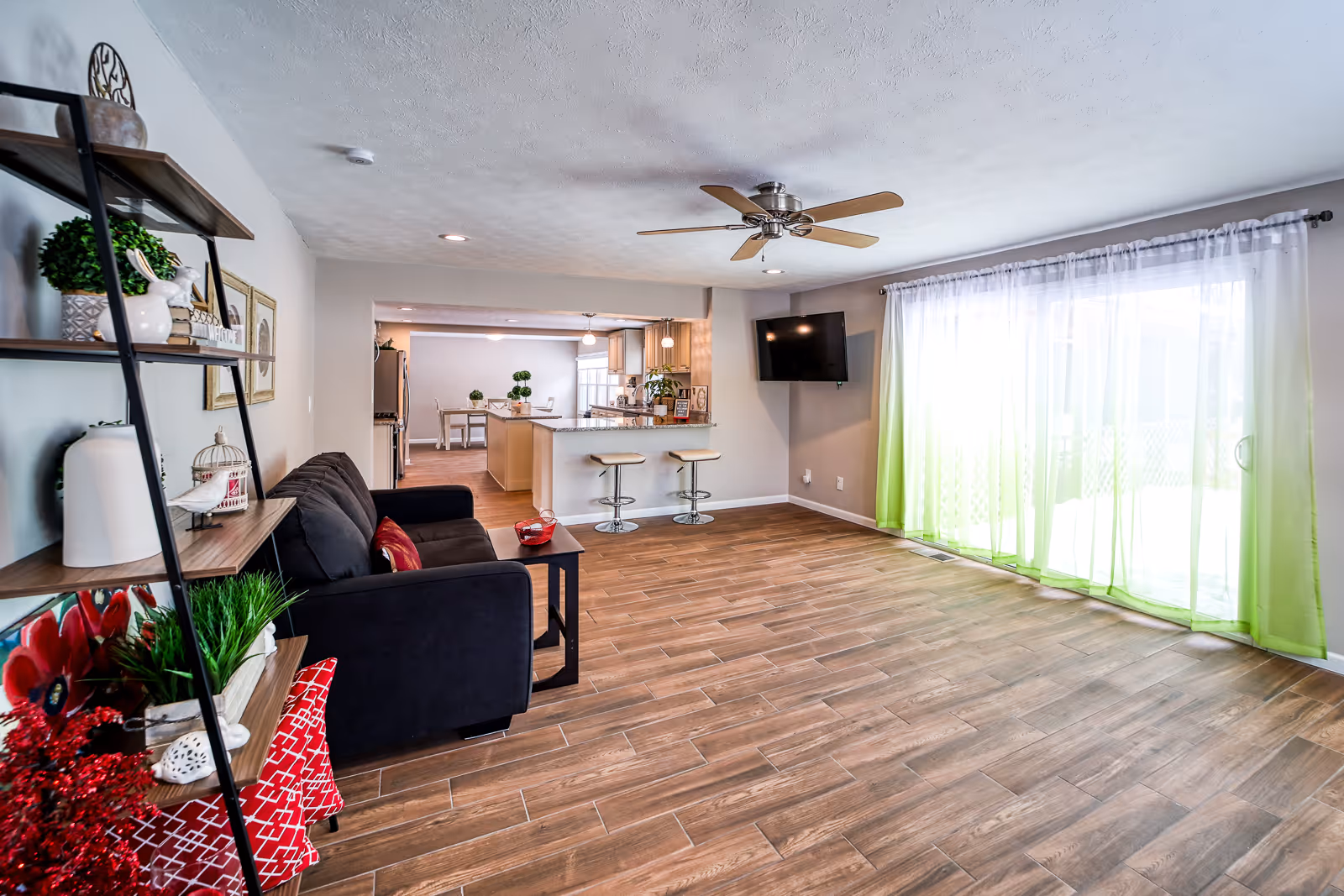 A bright and spacious living room with wooden flooring, a black sofa with red cushions, a black side table, and a black metal shelf with decorative items. The room has a ceiling fan, a wall-mounted TV, and large sliding glass doors with white and green curtains letting in natural light. In the background, there is a kitchen area with a breakfast bar and two bar stools, and a dining area with a table and chairs.