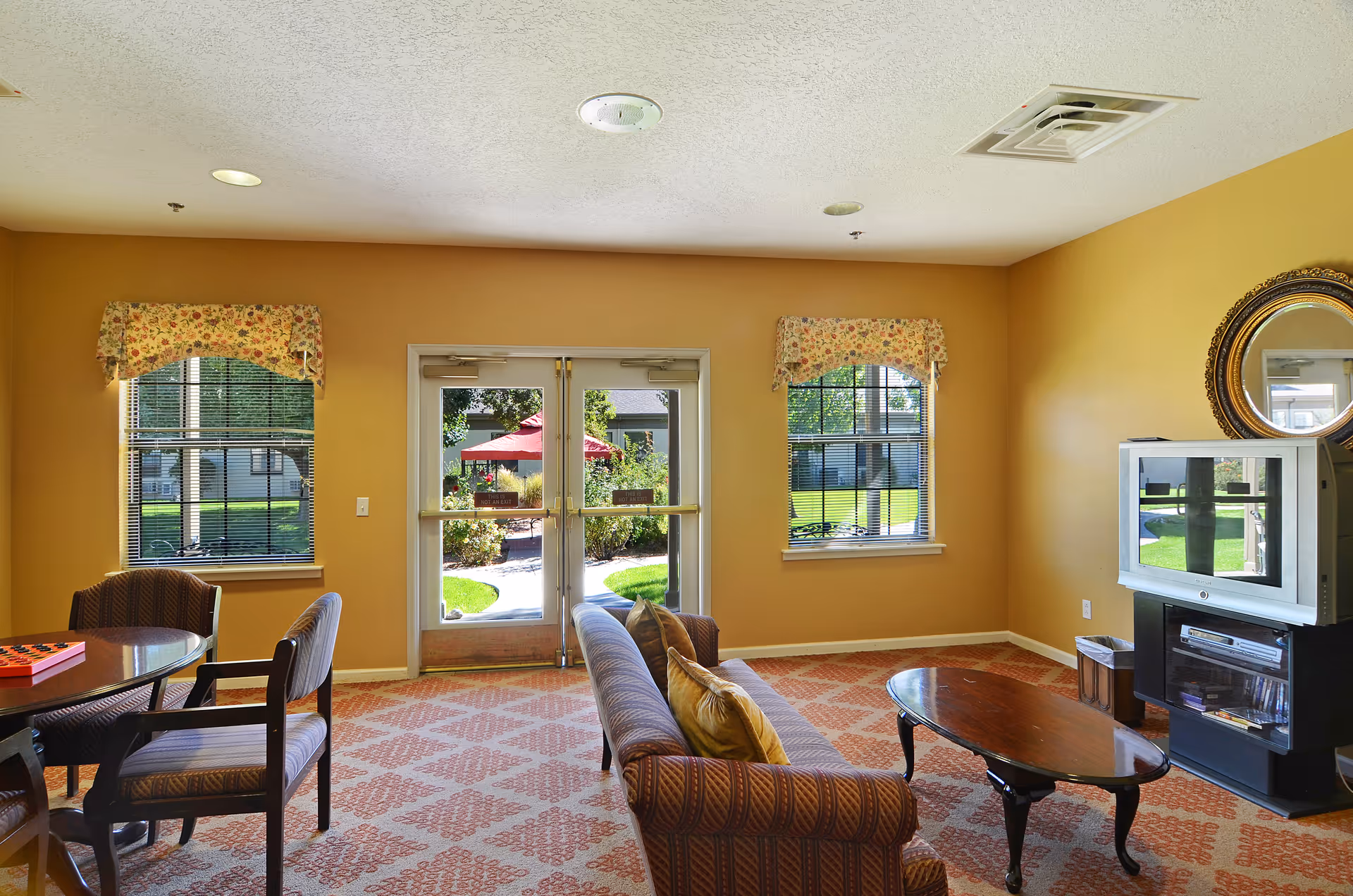 A cozy living room area with a patterned carpet, a sofa with cushions, a wooden coffee table, and a TV on a stand. There are two windows with floral valances and a glass door leading outside to a garden area with a red umbrella and greenery.