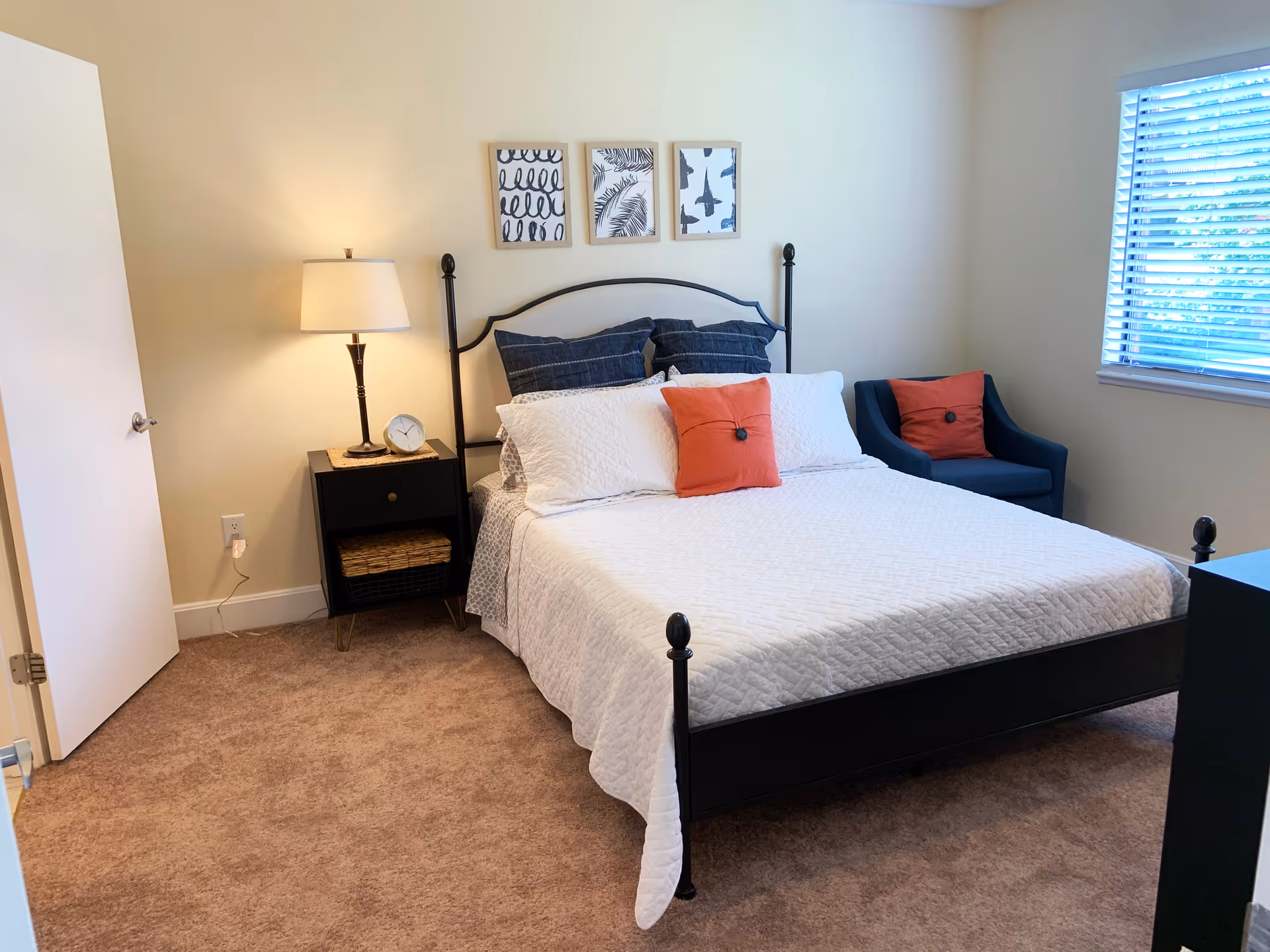 A neatly made bed with white quilted bedding and two orange accent pillows in a bedroom. The bed has a black metal frame with a white headboard. To the left of the bed is a black nightstand with a lamp and a clock on it. Above the bed are three framed black and white artworks. To the right of the bed is a blue armchair with an orange pillow. A window with white blinds lets in natural light on the right wall. The floor is carpeted in a light brown color.