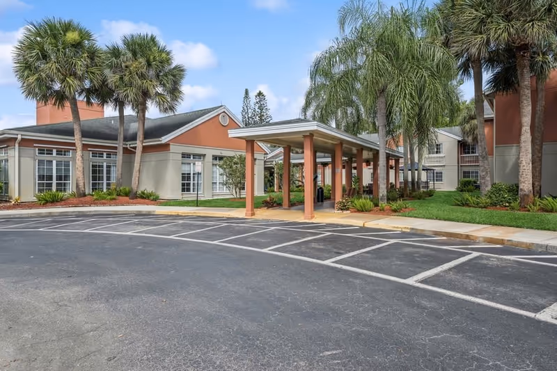 Exterior view of The Gardens of Port St. Lucie showing the entrance with a covered walkway, palm trees, and landscaped areas.