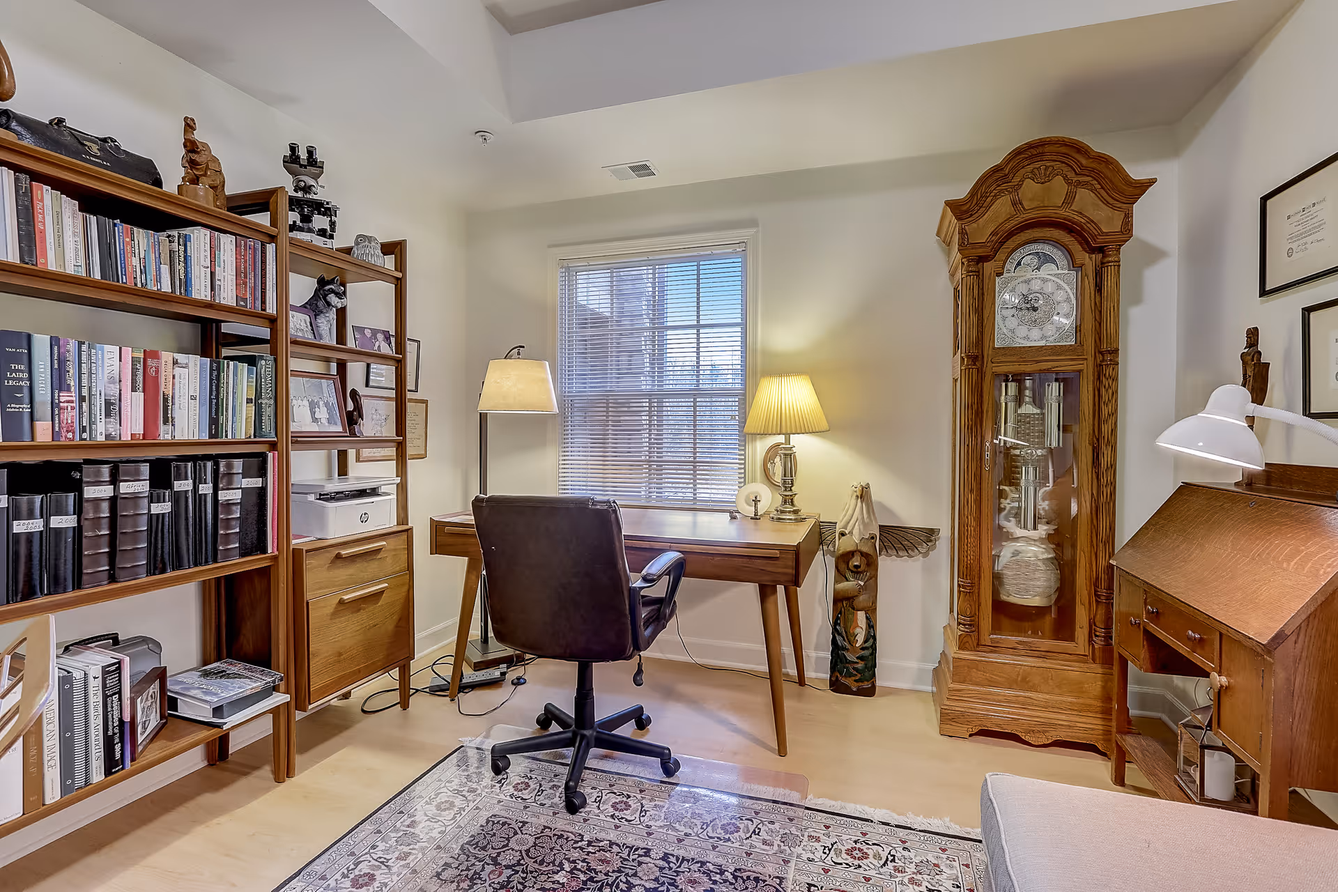 A cozy home office with a wooden desk and black swivel chair facing a window with blinds. To the left is a tall wooden bookshelf filled with books and binders. A floor lamp and a table lamp provide warm lighting. On the right side, there is a tall wooden grandfather clock and a wooden writing desk with a white desk lamp. A patterned rug covers part of the light wood floor.