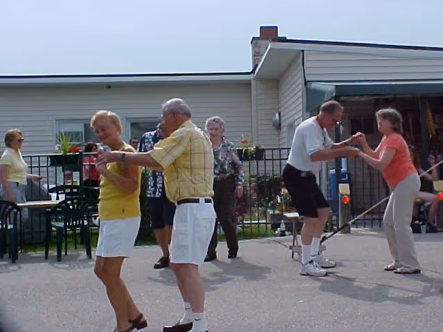 Several elderly people dancing and socializing outdoors on a paved area in front of a single-story building with light-colored siding. There are chairs and tables nearby, and some potted plants along a fence in the background.