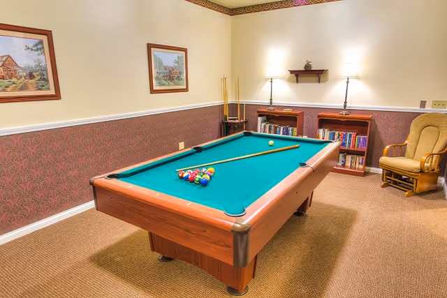 A cozy game room with a pool table in the center, set up with a cue stick and billiard balls. The room has patterned wallpaper with a chair rail, two framed paintings on the wall, a wooden rocking chair, two bookshelves filled with books, and two table lamps providing warm lighting.