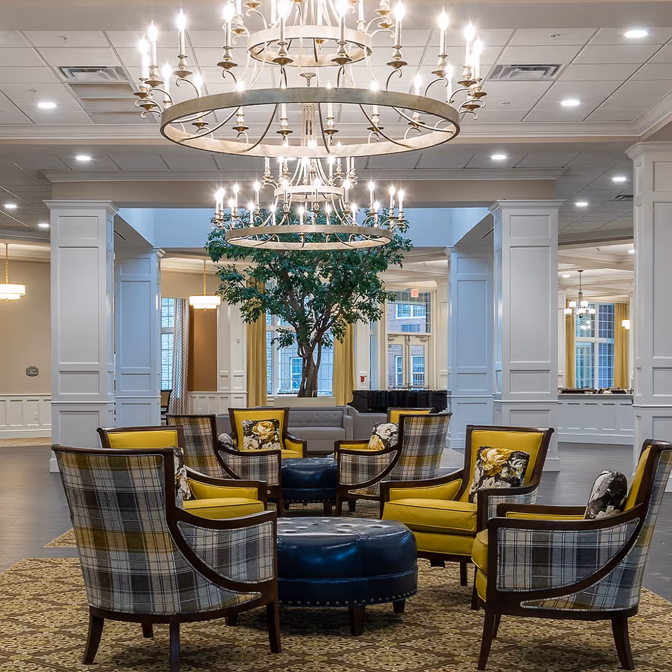 An elegant senior living lounge with circular chandeliers, upholstered chairs arranged around a blue ottoman and a large indoor tree.
