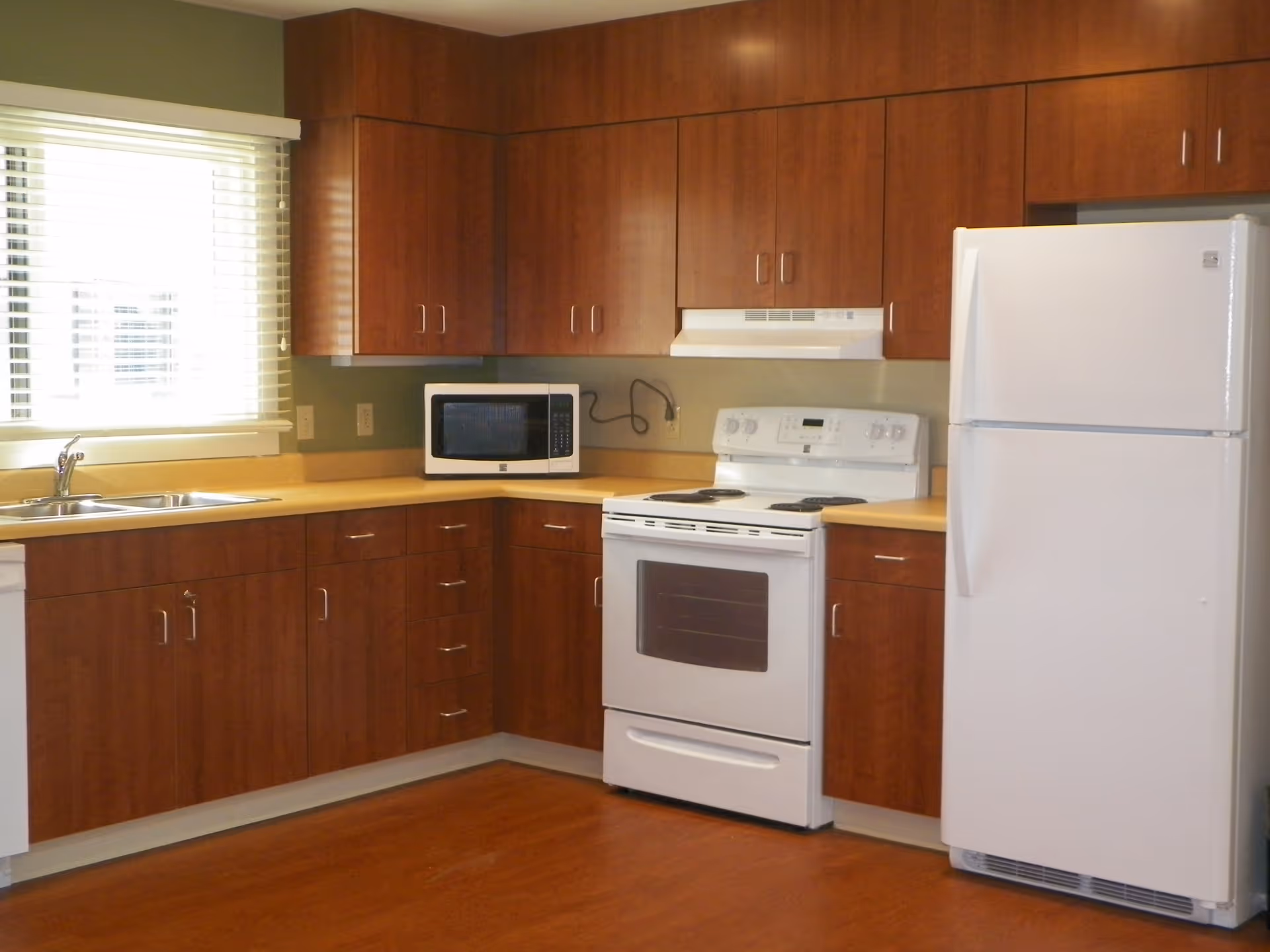 A kitchen with wooden cabinets, a white electric stove with oven, a white refrigerator, a microwave on the countertop, and a double sink under a window with blinds.