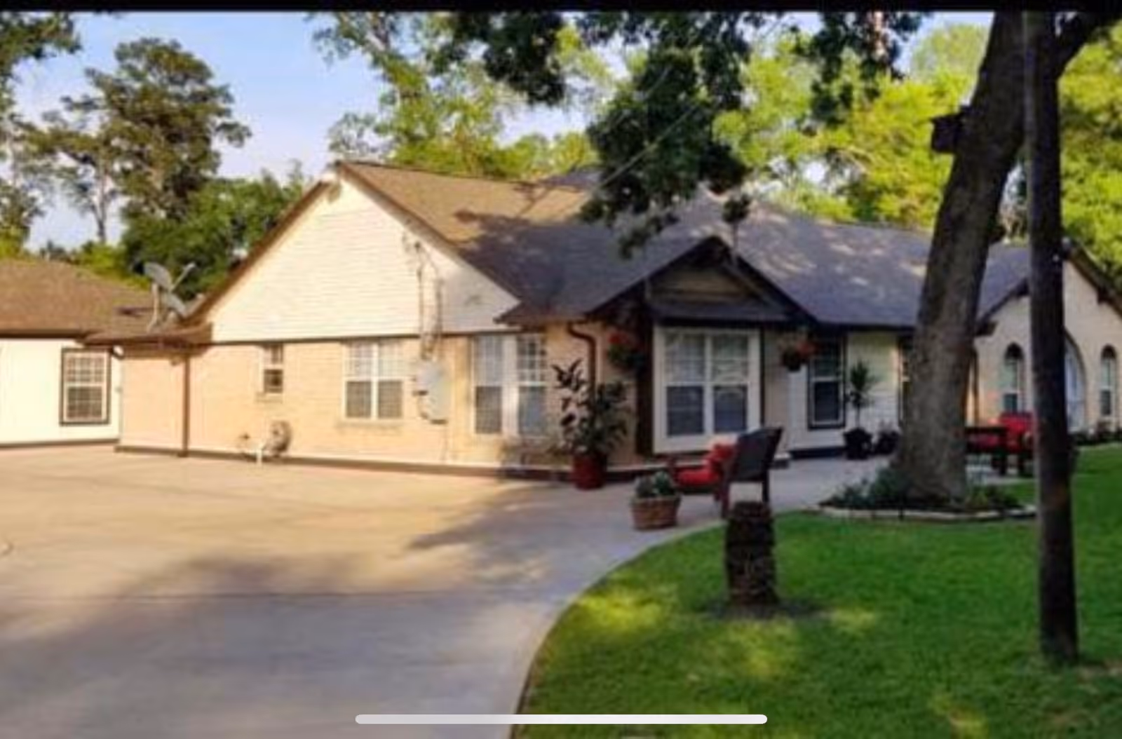 Exterior view of a single-story senior living facility building with beige walls and a brown roof. The building is surrounded by a paved driveway and green lawn with trees and outdoor seating areas featuring red cushions.