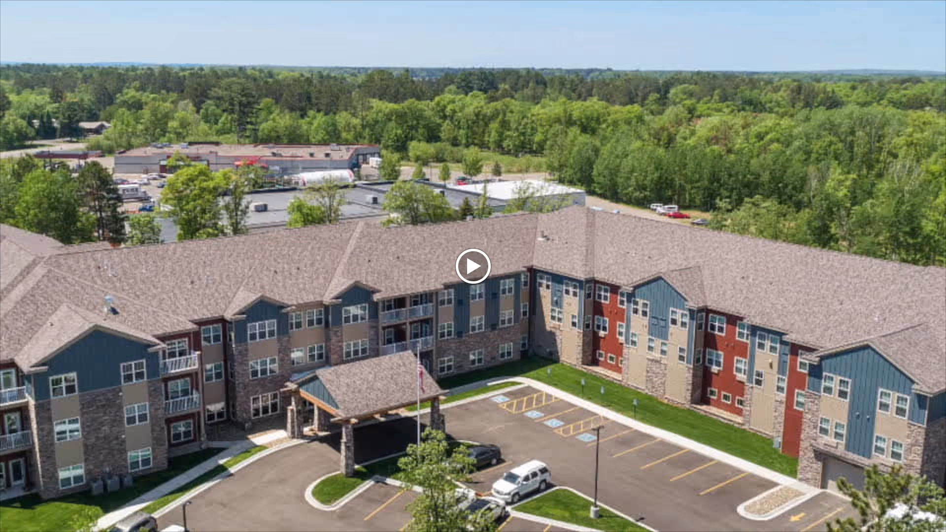 Aerial view of a large senior living facility building with a U-shaped layout, surrounded by greenery and parking spaces. The building has multiple floors with a mix of stone and siding exterior, and a covered entrance area with an American flag in front.