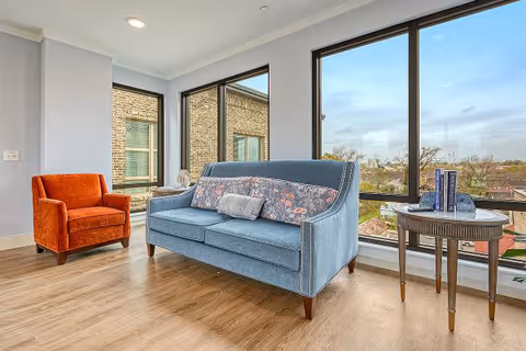 A bright living room area with large floor-to-ceiling windows showing an outside view. The room features a blue upholstered sofa with decorative pillows, an orange armchair, and a round wooden side table with books and decorative items on it. The floor is light wood, and the walls are painted light gray.