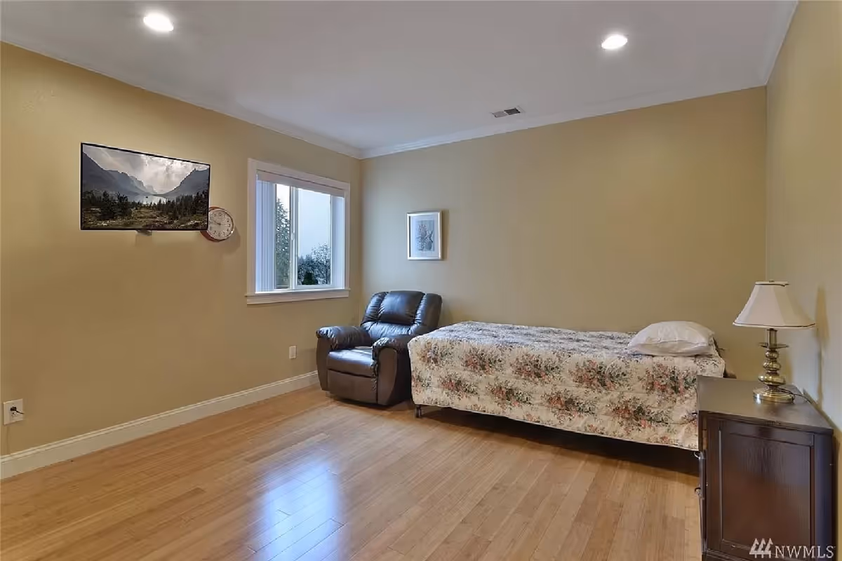 A simple bedroom with a single bed covered in a floral bedspread, a dark brown leather recliner chair next to a window, a small wooden nightstand with a lamp, a wall-mounted TV displaying a mountain landscape, and a clock on the wall.