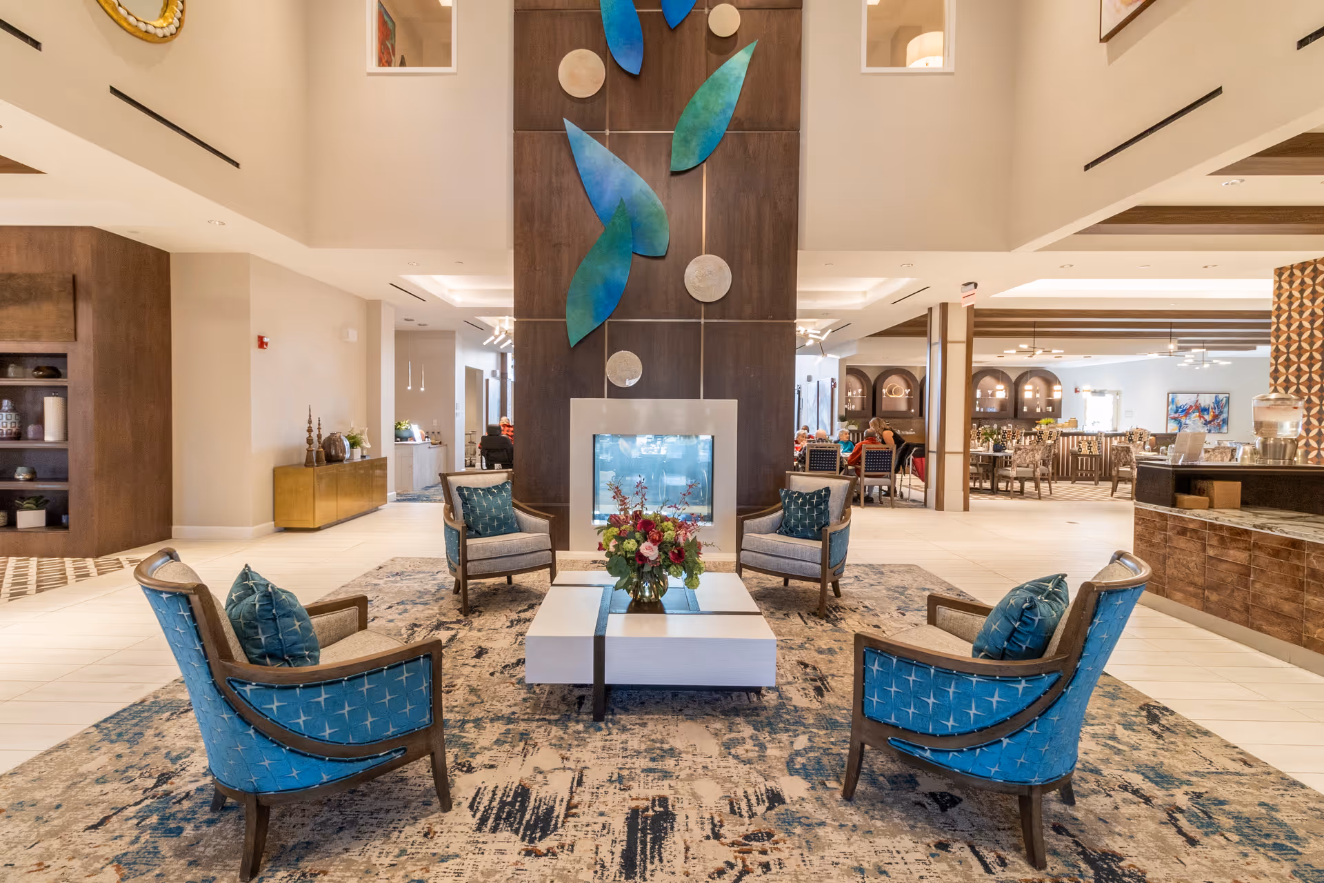 Bright modern senior living lobby with four blue chairs arranged around a central fireplace and a dining area visible in the background.